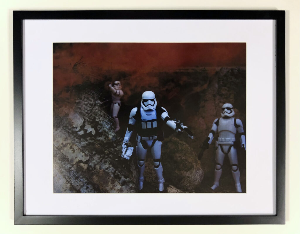 “Star Wars Edge” (2021) by Michael Aaron Gallagher. The 11” x 14” photograph was taken on June 27, 2021. Three different StormTroopers were used to create this toy photography scene. Front and center, is a 6” unbranded StormTrooper action figure with binoculars, imported from China. On the right, is a 6” Disney/Hasbro First Order StormTrooper. In the background, is a 3.5” Star Wars The Power of The Force Storm Trooper by Kenner. The smoke effect was created with a smoke ball from TNT Fireworks. The cave was created using the foundation of a demolished garage, a piece of broken concrete with wire mesh set at an angle, a piece of rotting bark under their feet and a rock.