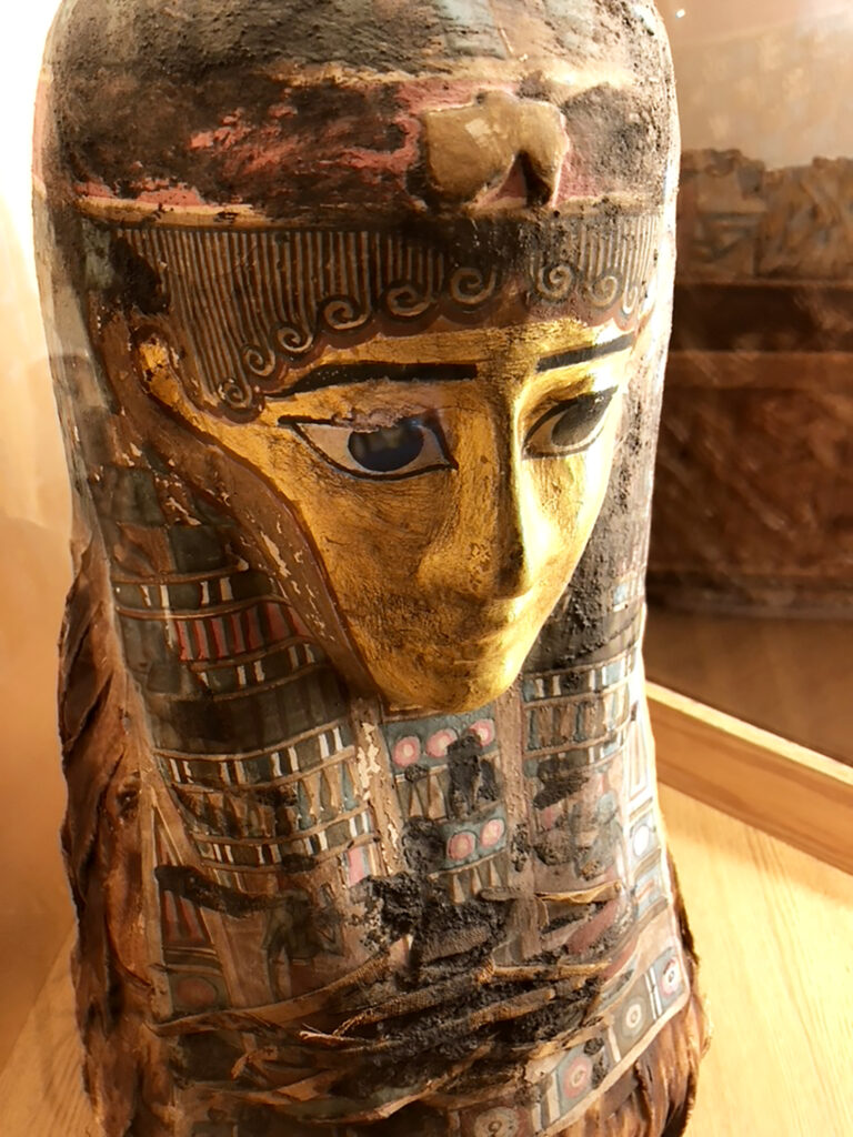 Egyptian mummy mask with mummy in the background at Cazenovia Library by Michael Aaron Gallagher